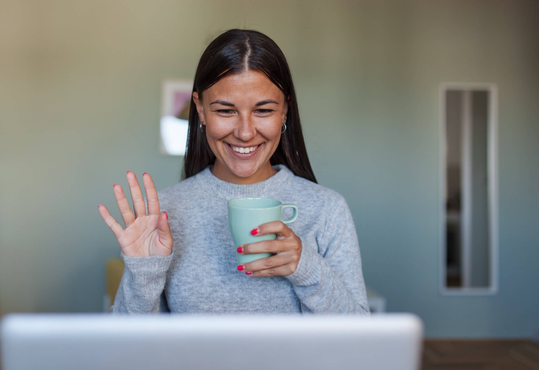 Woman holding mug and smiling and waving at laptop
