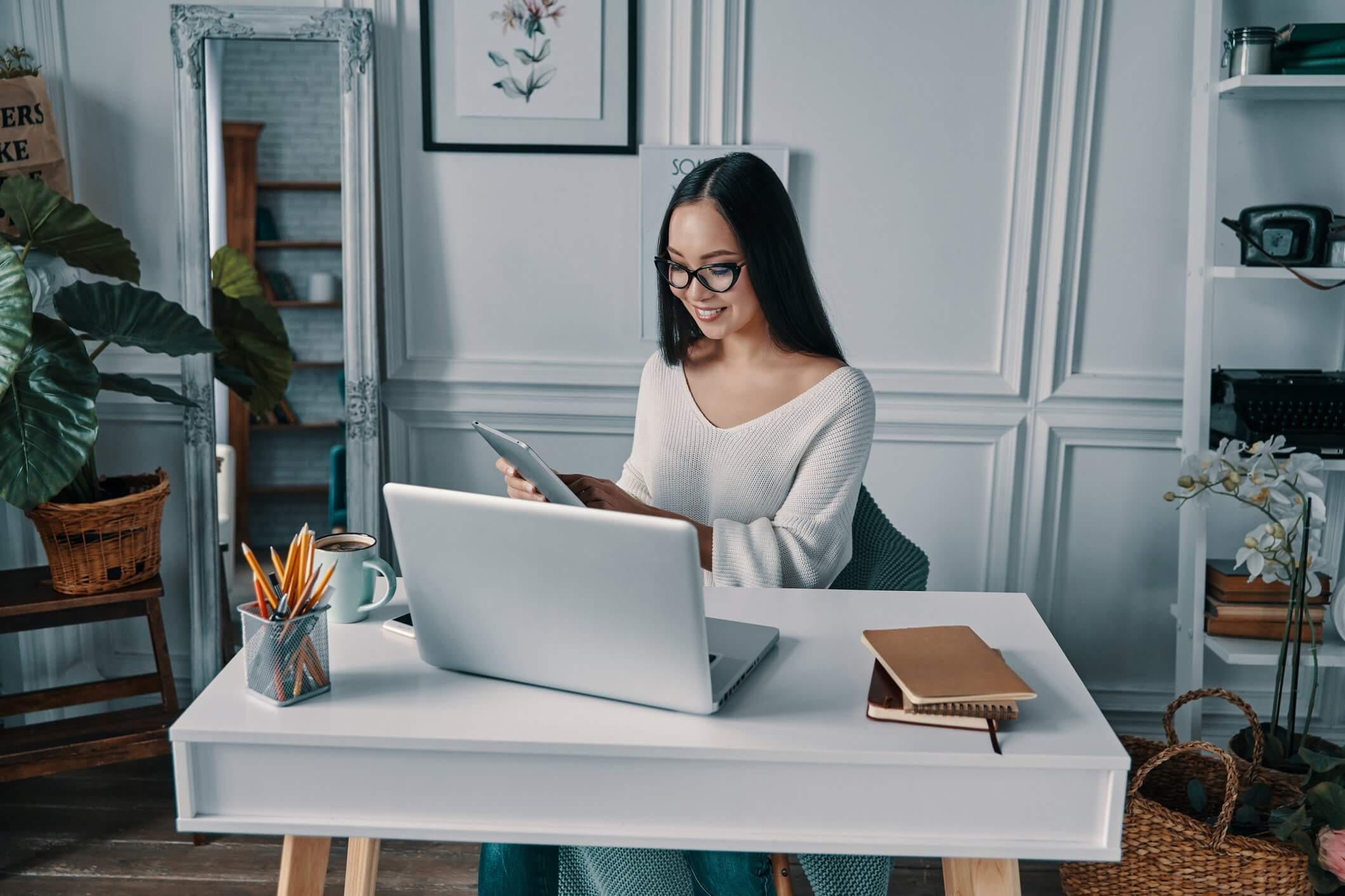 woman sitting at desk with notebooks and pencil holder and coffee cup and in front of laptop looking at phone