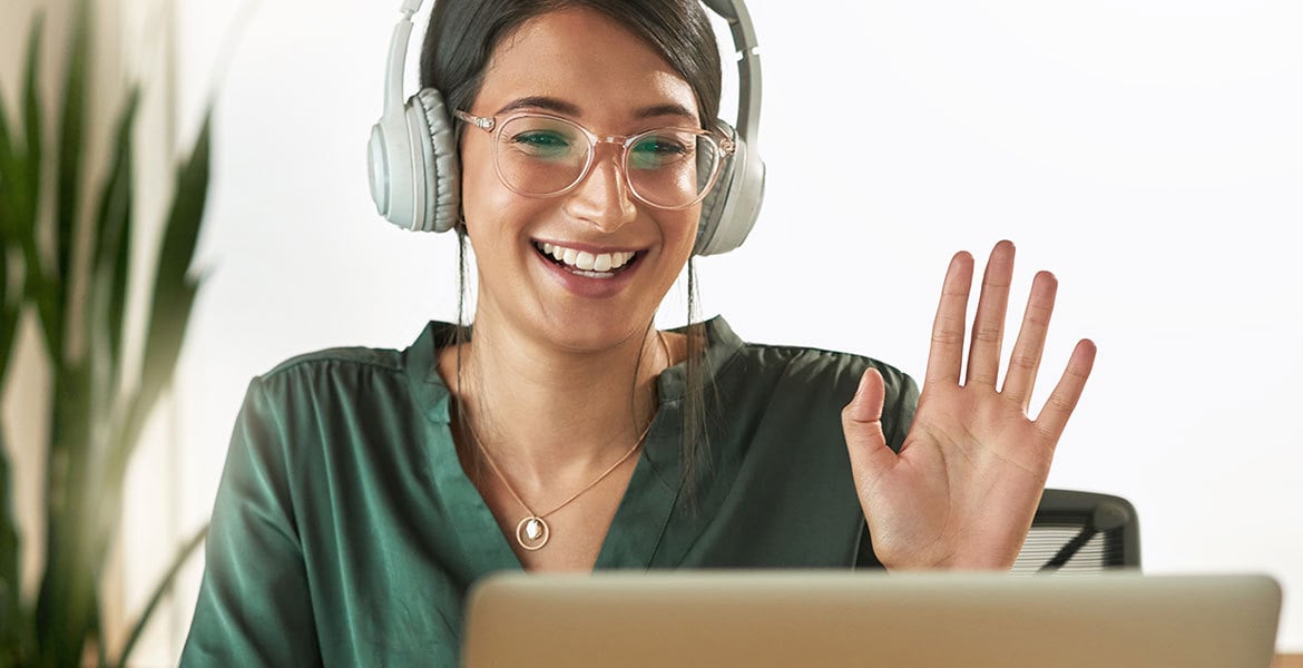 Young woman on computer wearing a headset, waving and smiling.
